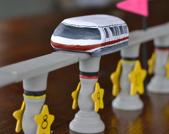 Könnte beinhalten: Ein weißer und roter Spielzeug-Monorail-Zug auf einem grauen Gleis mit gelben sternförmigen Sockeln mit den Nummern 8 und 9. Der Monorail befindet sich auf einer Holzoberfläche.