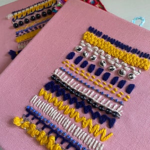 Op de afbeelding: Een roze canvas met een kleurrijk geometrisch borduurwerkontwerp. Het ontwerp bevat gele, witte, blauwe en zwarte draad en bevat kralen.