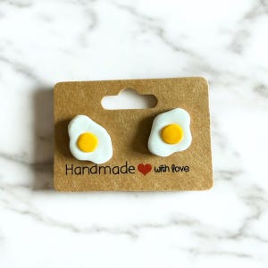 May include: A pair of handmade stud earrings in the shape of fried eggs, featuring a white egg white and a yellow yolk. The earrings are presented on a brown card with the text "Handmade with love".