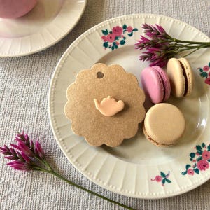 May include: A cream-coloured plate with a floral design, holding three macarons in pink and beige. A brown tag with a teapot design sits next to the macarons. A pink teacup is partially visible.