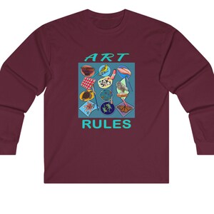 Pu&ograve; includere: Una t-shirt a maniche lunghe color granata con un design grafico di un quadrato blu che contiene varie immagini colorate. Il testo "ART RULES" &egrave; stampato in turchese sotto il quadrato.