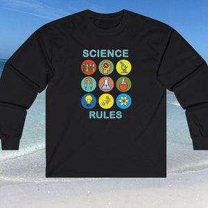 May include: Black long-sleeve shirt with a science theme. The shirt features the text 'SCIENCE RULES' in teal, with nine colorful science-related icons arranged in a grid above and below the text. The icons include test tubes, a gear, a brain, a microscope, a magnet, headphones, a flask, molecules, and an atom. The shirt is displayed against a beach background.