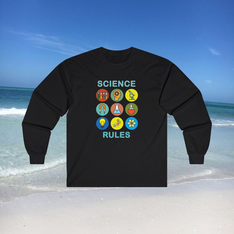 May include: Black long-sleeve shirt with a science theme. The shirt features the text 'SCIENCE RULES' in teal, with nine colorful science-related icons arranged in a grid above and below the text. The icons include test tubes, a gear, a brain, a microscope, a magnet, headphones, a flask, molecules, and an atom. The shirt is displayed against a beach background.