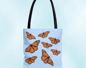 May include: A light blue tote bag with a black handle, featuring a pattern of orange and black monarch butterflies. The butterflies are in various sizes and orientations, creating a dynamic design. The bag is likely made of fabric.