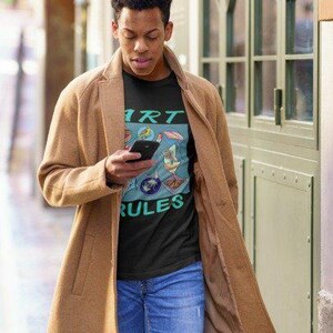 Pu&ograve; includere: Una persona indossa una maglietta nera con una grafica colorata che dice "Art Rules". La persona indossa un cappotto marrone e jeans blu.