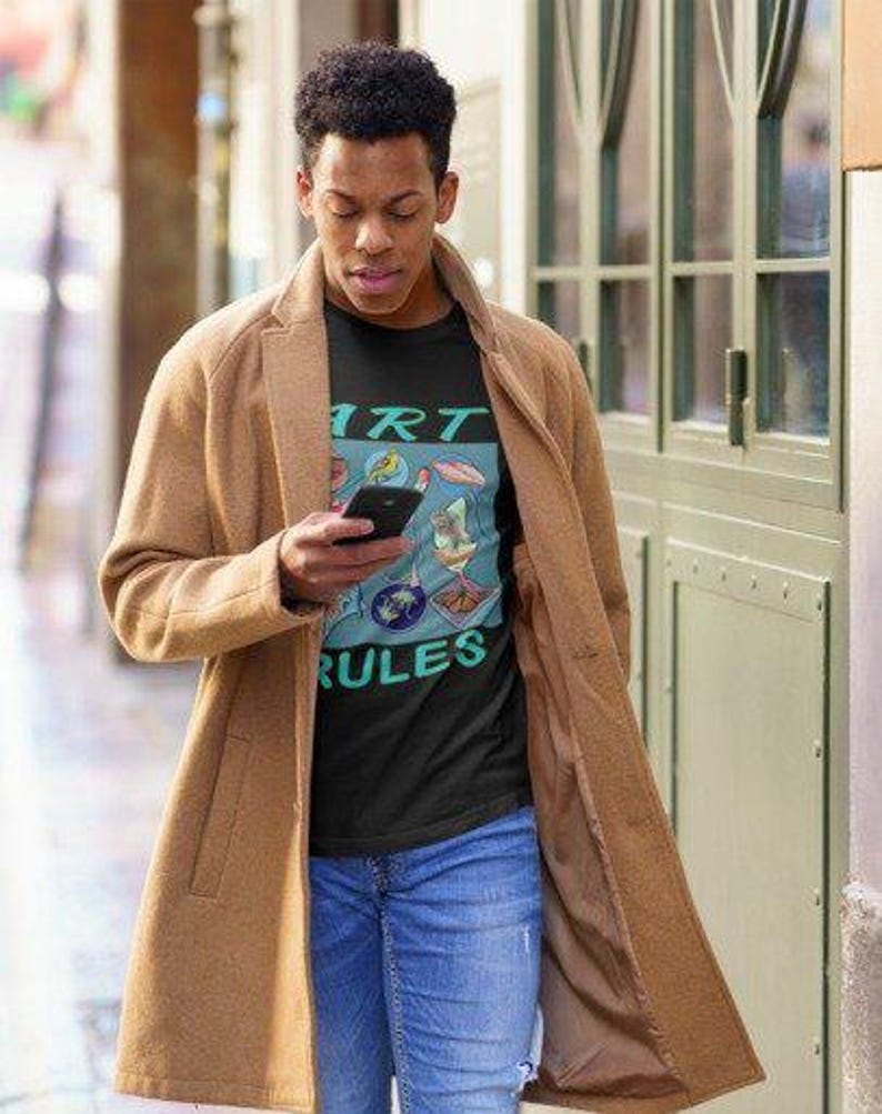Pu&ograve; includere: Una persona indossa una maglietta nera con una grafica colorata che dice "Art Rules". La persona indossa un cappotto marrone e jeans blu.