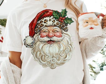 May include: White t-shirt featuring a Santa Claus graphic with a red hat, leopard print trim, and a long white beard. The Santa graphic is smiling. A Santa mug is held in the hand. The text "Design Barn Print Co." is in the bottom right corner.