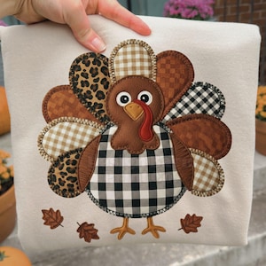 Könnte beinhalten: Ein cremefarbenes, quadratisches Kissen mit einem Truthahn-Applikation. Der Truthahn hat einen braunen Kopf und Körper, einen roten Wamm und ein schwarz-weiß kariertes Muster auf der Brust. Die Federn des Truthahns sind eine Mischung aus Mustern. Der Text "Design Barn Print Co." ist sichtbar.