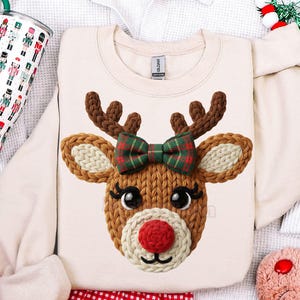 May include: Cream-colored sweatshirt featuring a knitted reindeer face with a red nose, brown antlers, and a green plaid bow. The sweatshirt is surrounded by a white waffle-knit fabric, a tumbler with a nutcracker design, and a red and white checkered fabric.