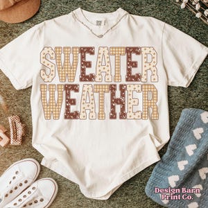 Puede incluir: Camiseta color crema con las palabras "SWEATER WEATHER" en un estampado a cuadros y floral marr&oacute;n y beige. La camiseta se combina con zapatillas blancas, un sombrero y una manta azul.
