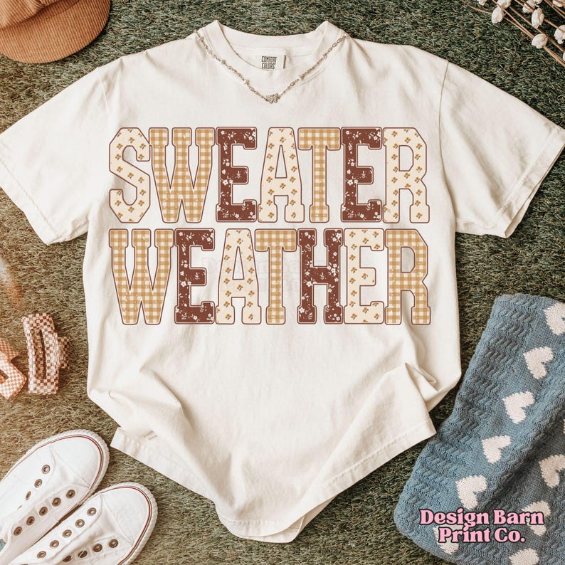 Puede incluir: Camiseta color crema con las palabras "SWEATER WEATHER" en un estampado a cuadros y floral marr&oacute;n y beige. La camiseta se combina con zapatillas blancas, un sombrero y una manta azul.
