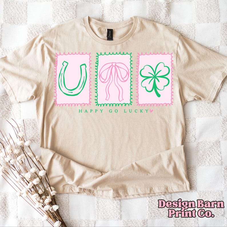 Pu&ograve; includere: T-shirt beige con un design grafico con un ferro di cavallo verde, un fiocco rosa e un trifoglio verde, ciascuno all'interno di una cornice smerlata rosa e verde. Il testo "HAPPY GO LUCKY" &egrave; in basso. La maglietta ha girocollo e maniche corte.