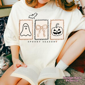 May include: Cream-colored t-shirt featuring a Halloween-themed design. The design includes a bat, a ghost, a bow, and a jack-o'-lantern, all within decorative frames. The text "SPOOKY SEASON" is printed below the frames.