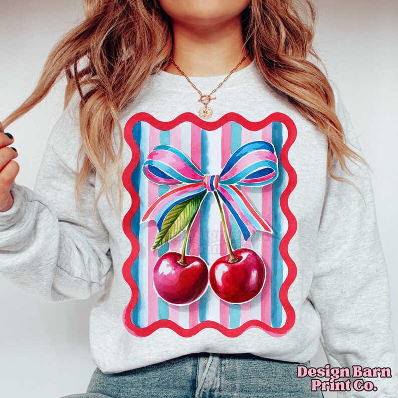 May include: A gray sweatshirt with a colorful graphic of two red cherries with a pink and blue bow on a striped background. The graphic is framed in a red scalloped border.