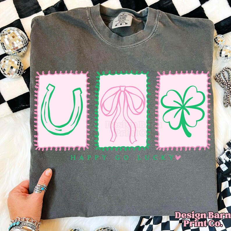 Pu&ograve; includere: T-shirt grigio scuro con tre disegni di francobolli rosa. Ogni francobollo presenta un ferro di cavallo verde, un fiocco rosa e un trifoglio verde. Il testo "HAPPY GO LUCKY" &egrave; stampato sotto. La maglietta &egrave; di Design Barn Print Co.