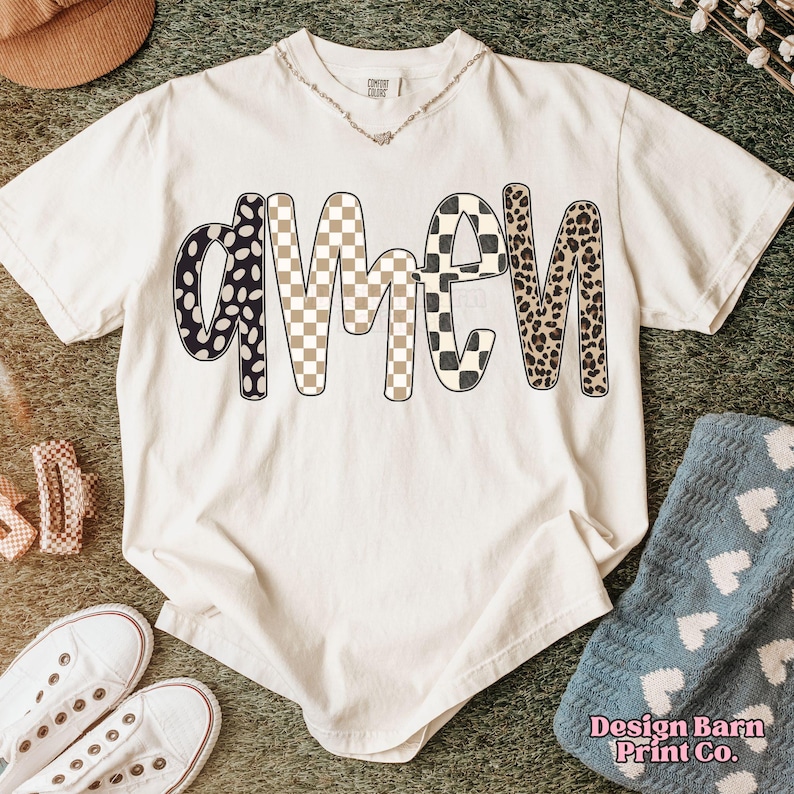 Op de afbeelding: Cr&egrave;mekleurig T-shirt met het woord "AMEN" in een opvallend patroon. De letters zijn voorzien van zwarte stippen, een beige en wit dambordpatroon, een zwart en wit dambordpatroon en een luipaardprint. Het shirt heeft een ronde hals en korte mouwen.