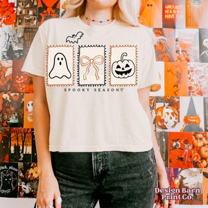 May include: Cream-colored t-shirt with a Halloween-themed design. The shirt features a bat, a ghost, a bow, and a jack-o'-lantern, all within decorative frames. The words "SPOOKY SEASON" are printed below the images.