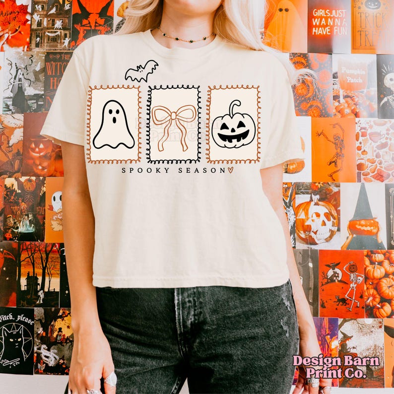 May include: Cream-colored t-shirt with a Halloween-themed design. The shirt features a bat, a ghost, a bow, and a jack-o'-lantern, all within decorative frames. The words "SPOOKY SEASON" are printed below the images.