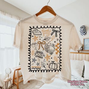 May include: Cream-colored t-shirt with a Thanksgiving-themed graphic. The design includes a pie, bow, turkey, stars, and the text "thanks giving" and "gobble til you wobble" within a black border. The shirt is hanging on a wooden hanger.