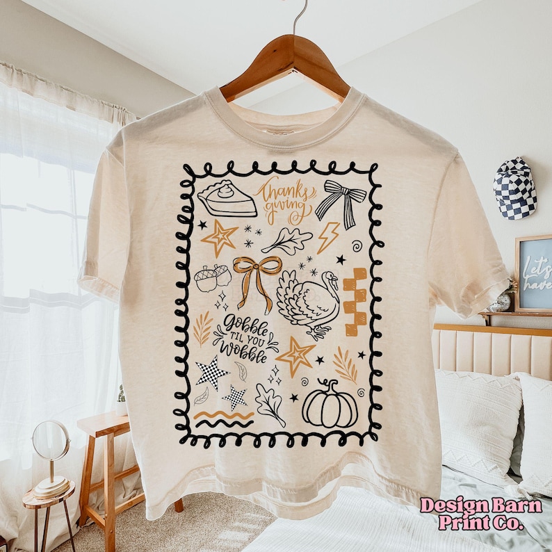 May include: Cream-colored t-shirt with a Thanksgiving-themed graphic. The design includes a pie, bow, turkey, stars, and the text "thanks giving" and "gobble til you wobble" within a black border. The shirt is hanging on a wooden hanger.