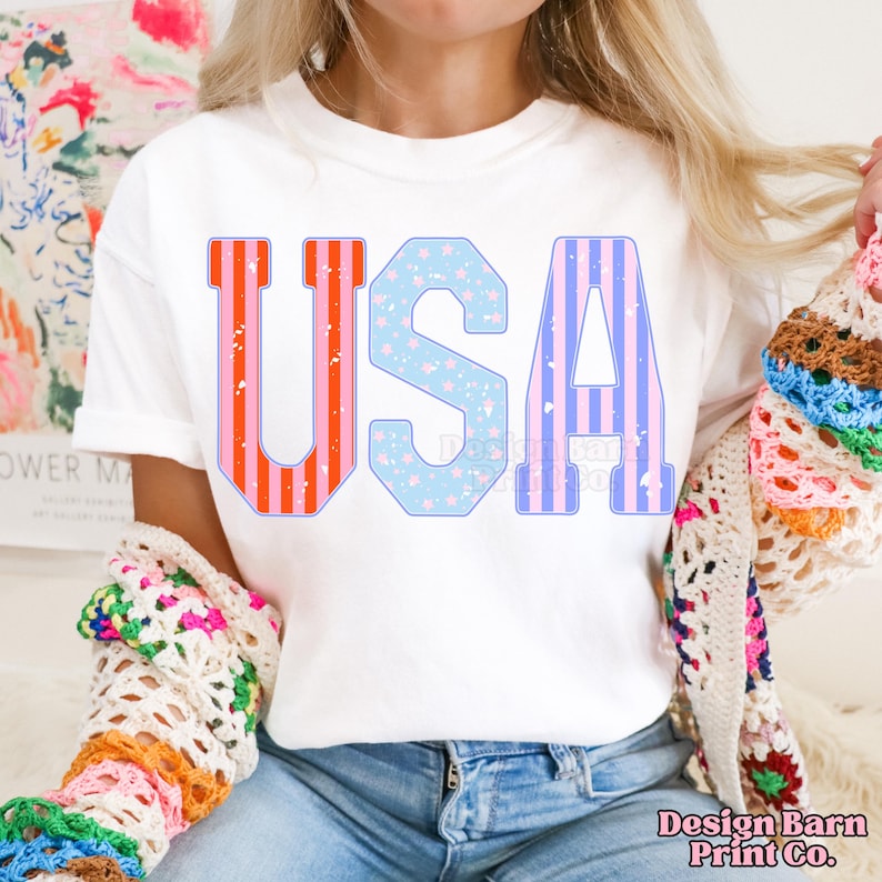 May include: A white t-shirt with the word "USA" printed on it in large, colorful letters. The letters are decorated with red, white, and blue stripes and stars.