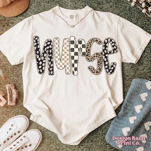 May include: Cream-colored t-shirt with the word "NURSE" in a bold, patterned design. The letters feature black and white polka dots, a tan and white checkerboard, and a leopard print. The shirt is styled with white sneakers and a necklace.