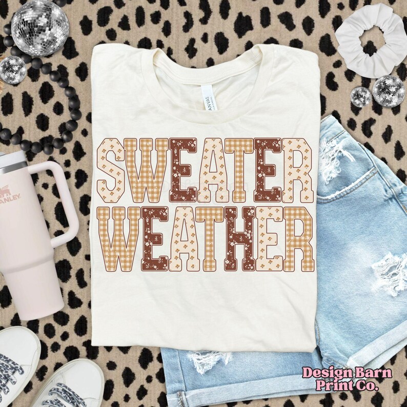 Puede incluir: Camiseta color crema con las palabras "SWEATER WEATHER" en un dise&ntilde;o estampado marr&oacute;n, beige y blanco. La camiseta se muestra con pantalones cortos de mezclilla y accesorios sobre un fondo de estampado de leopardo.