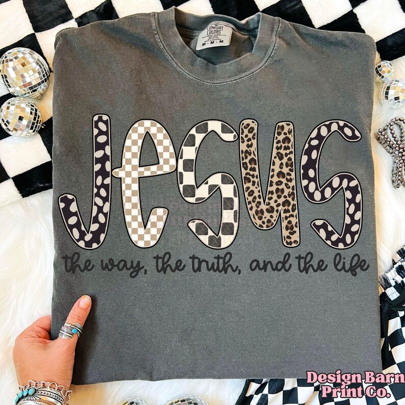 May include: Dark gray t-shirt with the word "JESUS" in a mix of patterns: polka dots, checkered, and leopard print. The text "the way, the truth, and the life" is printed below the word "JESUS".