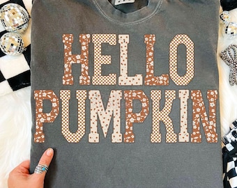 May include: A gray t-shirt with the words "HELLO PUMPKIN" in a mix of floral and checkered patterns. The shirt has a crew neck and short sleeves. The text is in shades of brown, cream, and white.