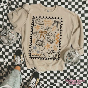 May include: Beige sweatshirt with a black and orange Thanksgiving-themed graphic. The design includes a turkey, pumpkin, pie, and the text "Gobble Til You Wobble" and "Thanksgiving". The sweatshirt is on a black and white checkered surface.