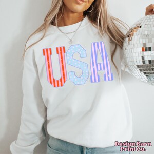 May include: White sweatshirt with the word "USA" printed in red, white, and blue stripes and stars. The letters are outlined in blue.