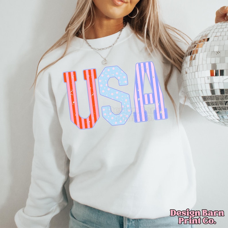 May include: White sweatshirt with the word "USA" printed in red, white, and blue stripes and stars. The letters are outlined in blue.