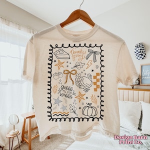 Puede incluir: Camiseta color crema con un dise&ntilde;o tem&aacute;tico de Acci&oacute;n de Gracias. El dise&ntilde;o incluye un pastel, un lazo, un pavo, estrellas y los textos "thanks giving" y "gobble til you wobble" dentro de un borde negro. La camiseta est&aacute; colgada en una percha de madera.