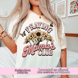 May include: A white t-shirt featuring a pink and yellow graphic design that reads "Praying Mama's Club". The design incorporates sunflowers and butterflies.