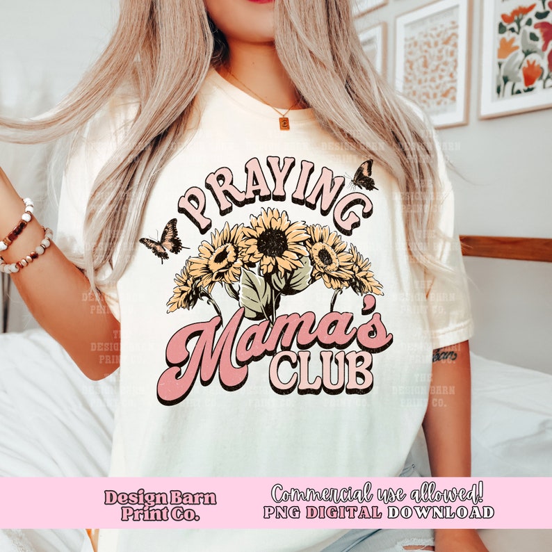 May include: A white t-shirt featuring a pink and yellow graphic design that reads "Praying Mama's Club". The design incorporates sunflowers and butterflies.