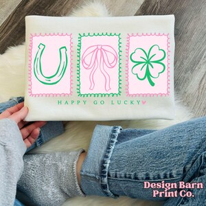 Pu&ograve; includere: Felpa color crema con tre disegni grafici rosa e verdi. I disegni includono un ferro di cavallo, un fiocco e un quadrifoglio. Il testo "HAPPY GO LUCKY" &egrave; stampato sotto i disegni.