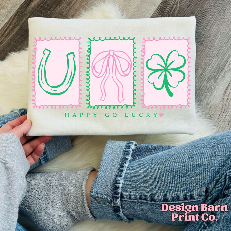 Pu&ograve; includere: Felpa color crema con tre disegni grafici rosa e verdi. I disegni includono un ferro di cavallo, un fiocco e un quadrifoglio. Il testo "HAPPY GO LUCKY" &egrave; stampato sotto i disegni.