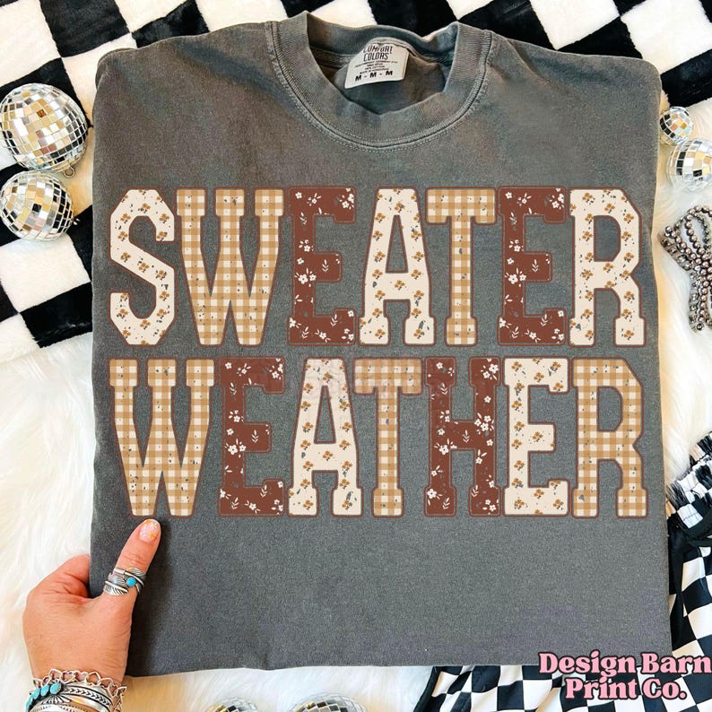 Puede incluir: Una camiseta gris con las palabras "SWEATER WEATHER" en letras grandes y decorativas. Las letras est&aacute;n rellenas de patrones florales y a cuadros en tonos marrones y beige. La camiseta tiene cuello redondo.