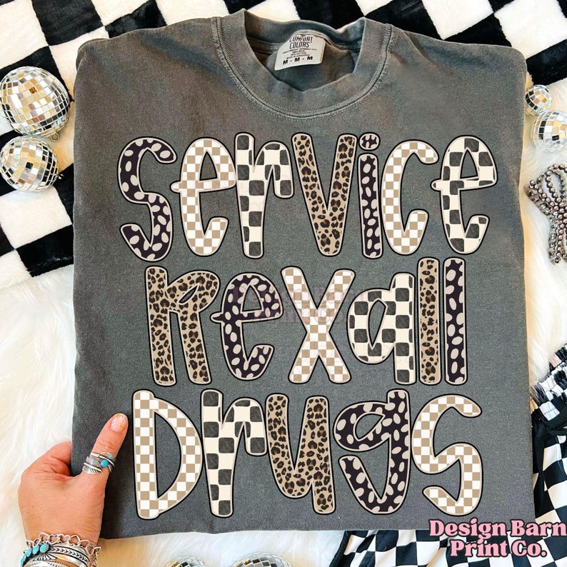 May include: Dark gray t-shirt with the words "SERVICE REXALL DRUGS" in a mix of checkered and leopard print lettering. The shirt is a Comfort Colors brand and has a crew neck.