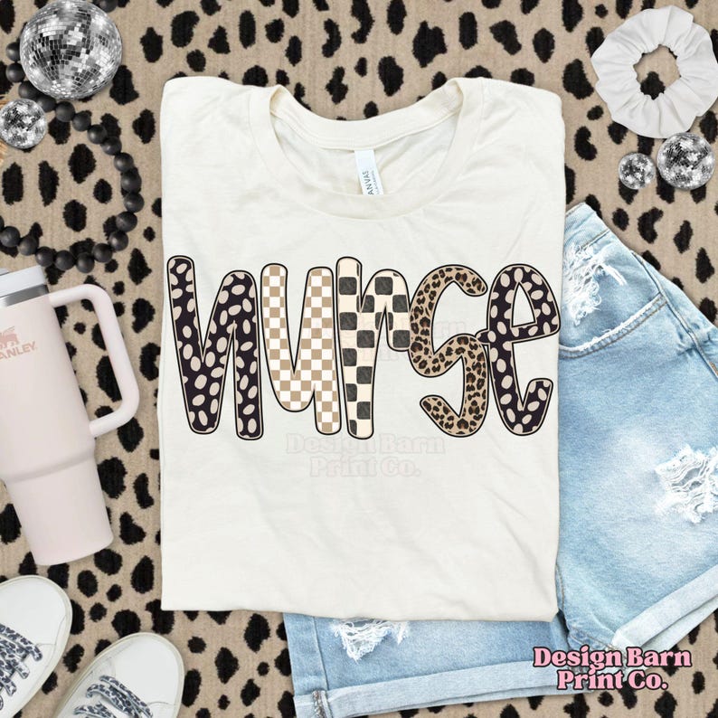 May include: Cream-colored t-shirt with the word "NURSE" in a bold, patterned design. The letters feature a mix of black and white polka dots, tan and white checkered, and leopard print patterns. The shirt is paired with denim shorts.