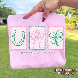 Pu&ograve; includere: Una felpa rosa chiaro con tre grafiche incorniciate: un ferro di cavallo verde, un fiocco rosa e un quadrifoglio verde. Il testo "HAPPY GO LUCKY" &egrave; sotto le grafiche. Il logo Design Barn Print Co. &egrave; in basso a destra.
