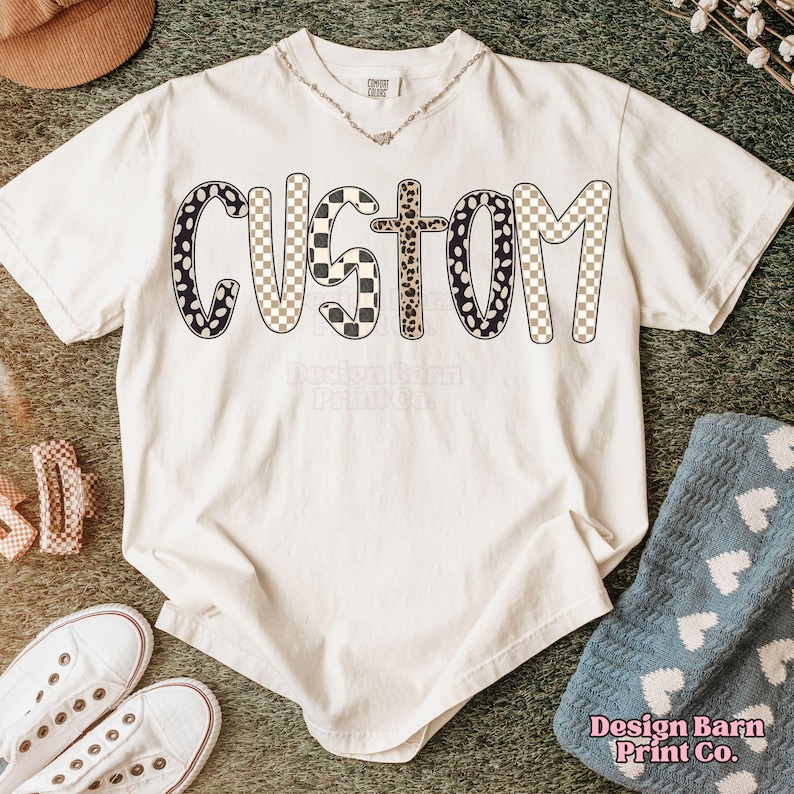 May include: Cream-colored t-shirt with the word "CUSTOM" in a bold, patterned design. The letters feature black and white polka dots, checkered patterns, and a leopard print. The shirt is styled with white sneakers and a blue blanket.