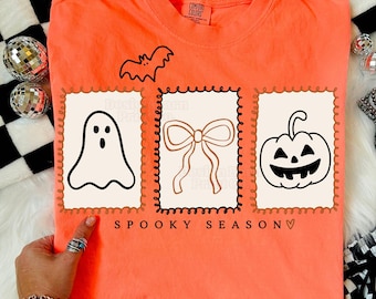 May include: Orange t-shirt with a bat graphic and three framed illustrations: a ghost, a bow, and a jack-o'-lantern. The text "Spooky Season" is printed below the illustrations. The shirt is a Comfort Colors brand.