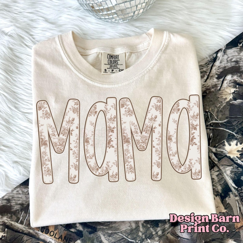 May include: Cream-colored t-shirt with the word "MAMA" in large, floral-patterned letters. The shirt is folded neatly, with the brand tag visible at the collar. The text "Design Barn Print Co." is printed in the bottom right corner.