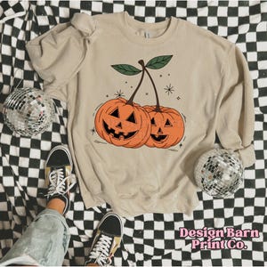 K&ouml;nnte beinhalten: Ein beiger Pullover mit einem Aufdruck von zwei orangefarbenen K&uuml;rbissen mit Jack-o'-Lantern-Gesichtern auf einem Stiel. Die K&uuml;rbisse sind so gestaltet, dass sie wie Kirschen aussehen.