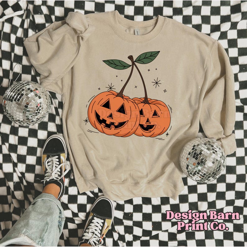K&ouml;nnte beinhalten: Ein beiger Pullover mit einem Aufdruck von zwei orangefarbenen K&uuml;rbissen mit Jack-o'-Lantern-Gesichtern auf einem Stiel. Die K&uuml;rbisse sind so gestaltet, dass sie wie Kirschen aussehen.