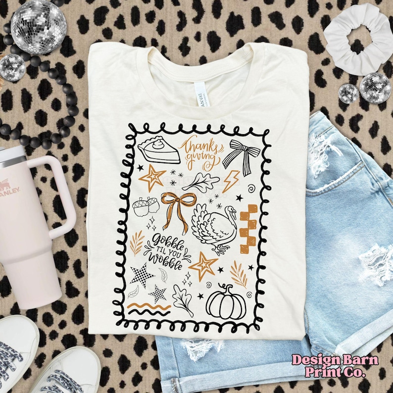 Puede incluir: Camiseta color crema con un dise&ntilde;o tem&aacute;tico de Acci&oacute;n de Gracias. El dise&ntilde;o incluye una porci&oacute;n de pastel, un pavo, una calabaza y los textos "Thanksgiving" y "Gobble Til You Wobble". El gr&aacute;fico est&aacute; delineado en negro y presenta acentos marrones y dorados.