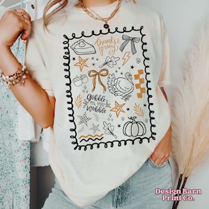 Puede incluir: Camiseta color crema con un dise&ntilde;o tem&aacute;tico de Acci&oacute;n de Gracias. El dise&ntilde;o incluye una porci&oacute;n de pastel, un lazo, un pavo, estrellas y los textos "Thanksgiving" y "Gobble Til You Wobble" en negro, marr&oacute;n y dorado.