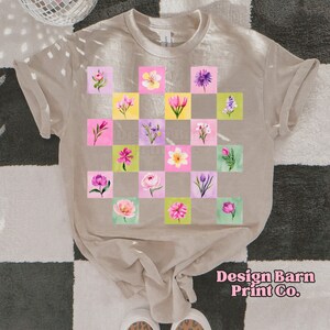 May include: Beige t-shirt featuring a checkerboard pattern of watercolor flowers in pink, yellow, and purple squares. The shirt has rolled-up sleeves and the text "Design Barn Print Co." in pink script.