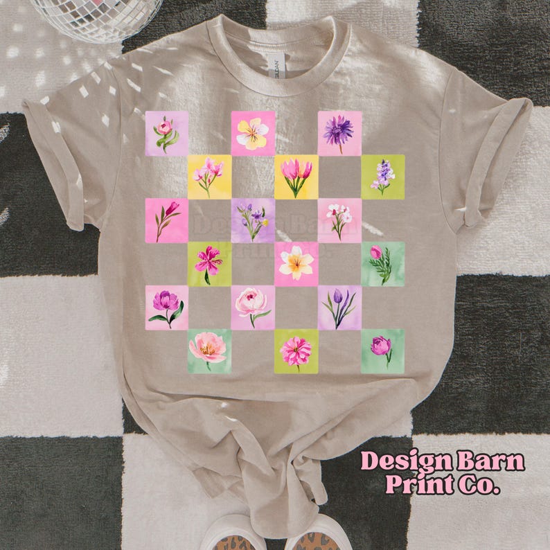 May include: Beige t-shirt featuring a checkerboard pattern of watercolor flowers in pink, yellow, and purple squares. The shirt has rolled-up sleeves and the text "Design Barn Print Co." in pink script.